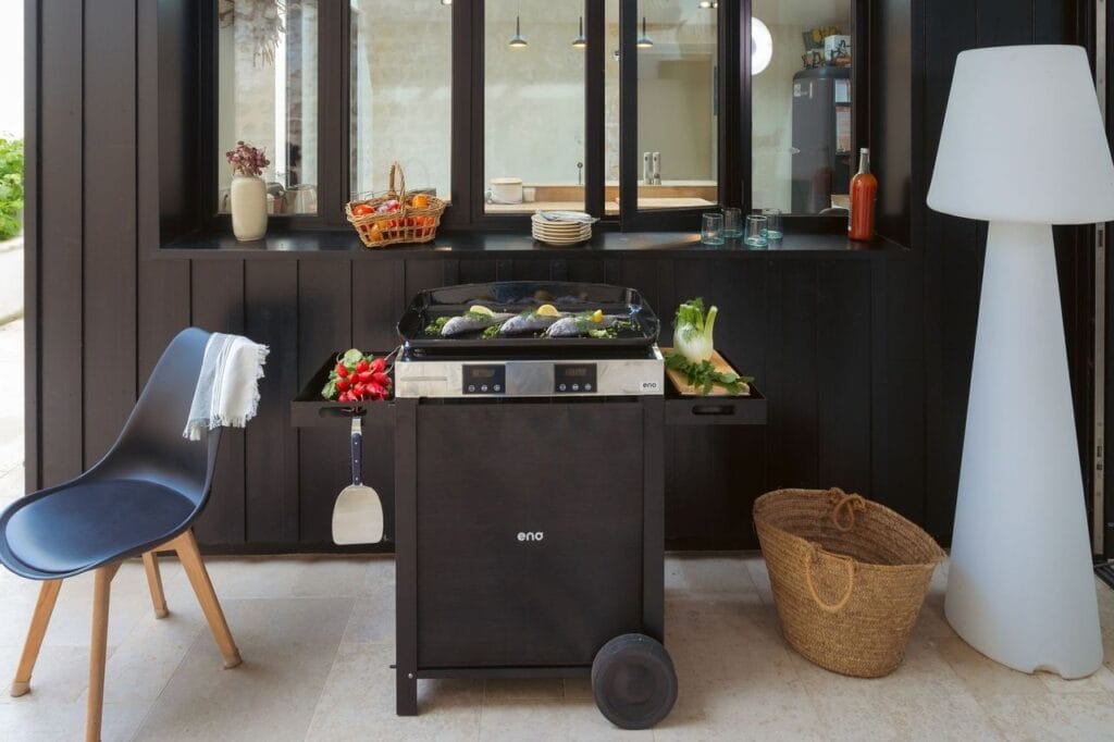Plancha électrique installée sur une terrasse en pierre naturelle devant une façade en bois noir, éclairée par une lampe design blanche.