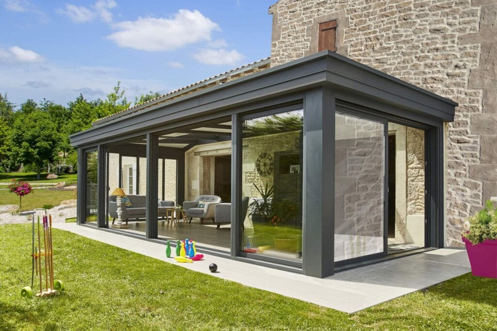 Pergola vitrée Concept Alu en aluminium noir avec salon de jardin sur terrasse composite, haie taillée et éclairage LED intégré