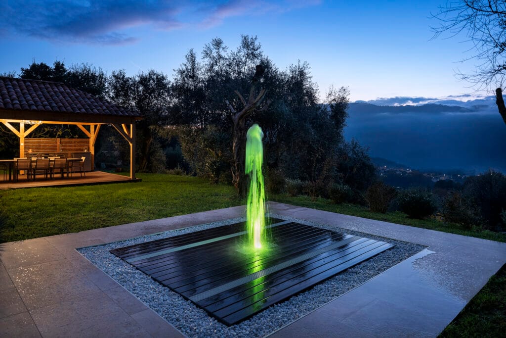 Jet d'eau Deckflow illuminé en vert fluo la nuit sur une terrasse en bois, avec un pool house en arrière-plan