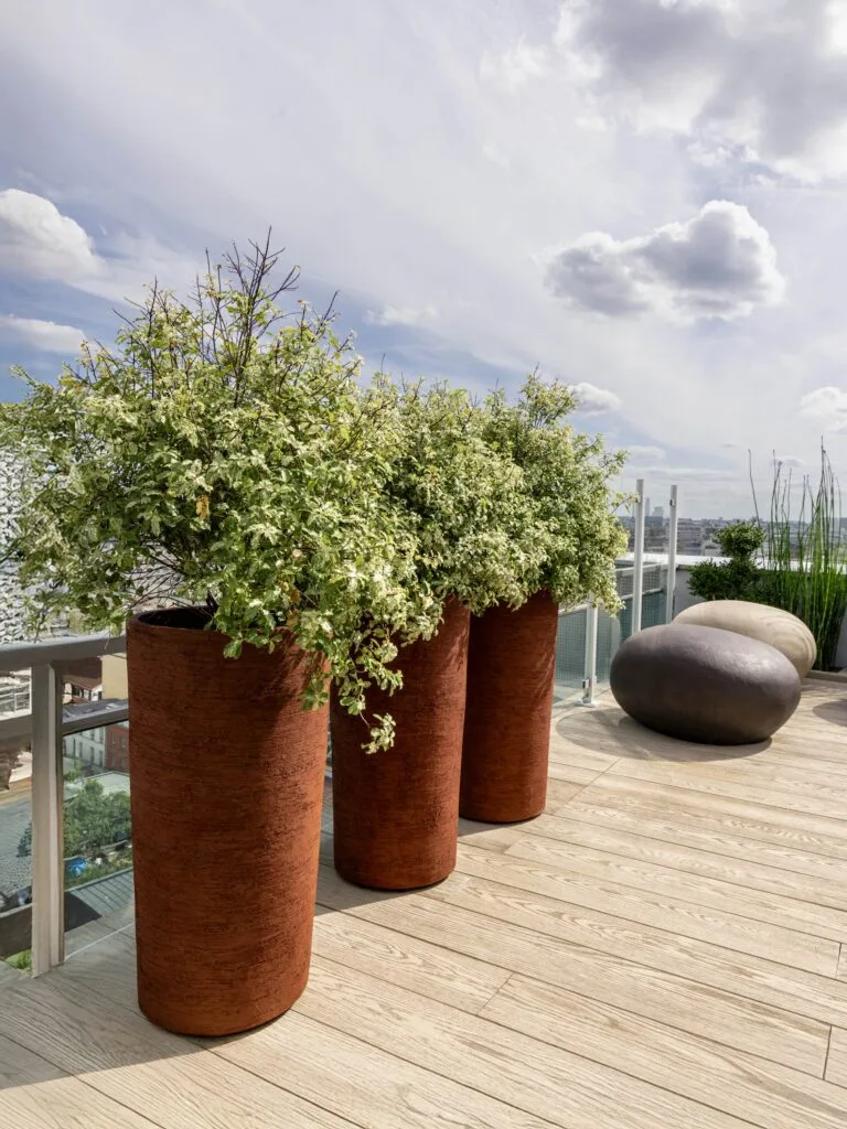 Trois grands pots de fleurs Vierkant cylindriques couleur terre cuite rouille sur une terrasse sur le toit avec vue, sol en bois clair et galets décoratifs noirs