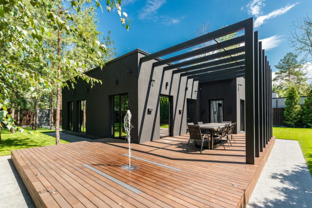 Grande terrasse en bois sous pergola noire moderne animée par un jet d'eau central Deckflow devant une maison contemporaine