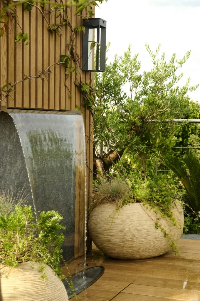 Fontaine cascade en métal et grand pot de jardin texturé couleur crème planté de verdure, sur une terrasse en bois