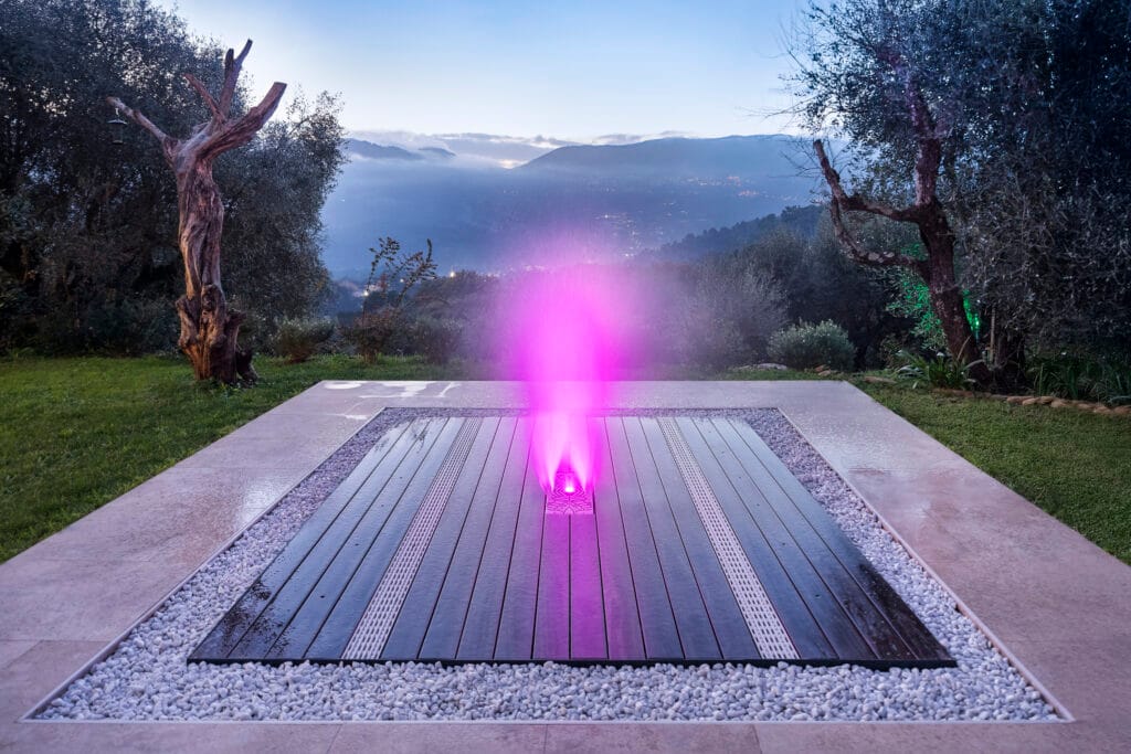 Vue nocturne d'une fontaine de terrasse Deckflow rétroéclairée par LED rose, entourée de galets blancs