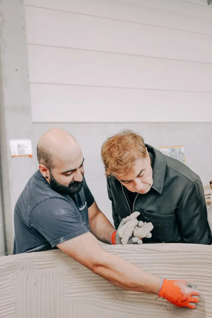 César Saint-ouen et un artisan (un barbu, un plus âgé) travaillant ensemble sur l'application de la matière sur un grand pot de jardin en terre cuite, gestes précis